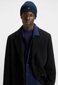 Navy ribbed knit beanie with logo tab; paired with a black wool coat and navy button-up shirt. Modern, structured fit, minimal detailing.