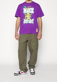 Fialové grafické tričko s textem "Nike Just Do It" a designem cheruba. V kombinaci s olivovými cargo kalhotami a černými, bílými a červenými teniskami.