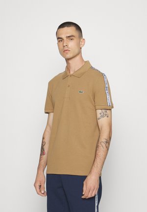 Camisa polo marrón de manga corta, con tapeta de tres botones y logotipo verde; presenta acentos a rayas blancas en los hombros.