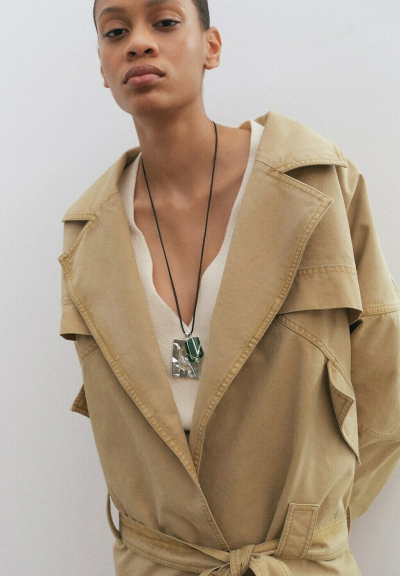 Femme portant un trench-coat beige par-dessus un haut blanc, avec un collier pendentif vert et argenté, sur un fond clair uni.