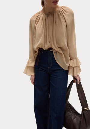 Beige Faltenbluse mit langen, rüschigen Ärmeln, kombiniert mit hoch taillierten dunkelblauen Jeans und einer braunen Ledertasche.