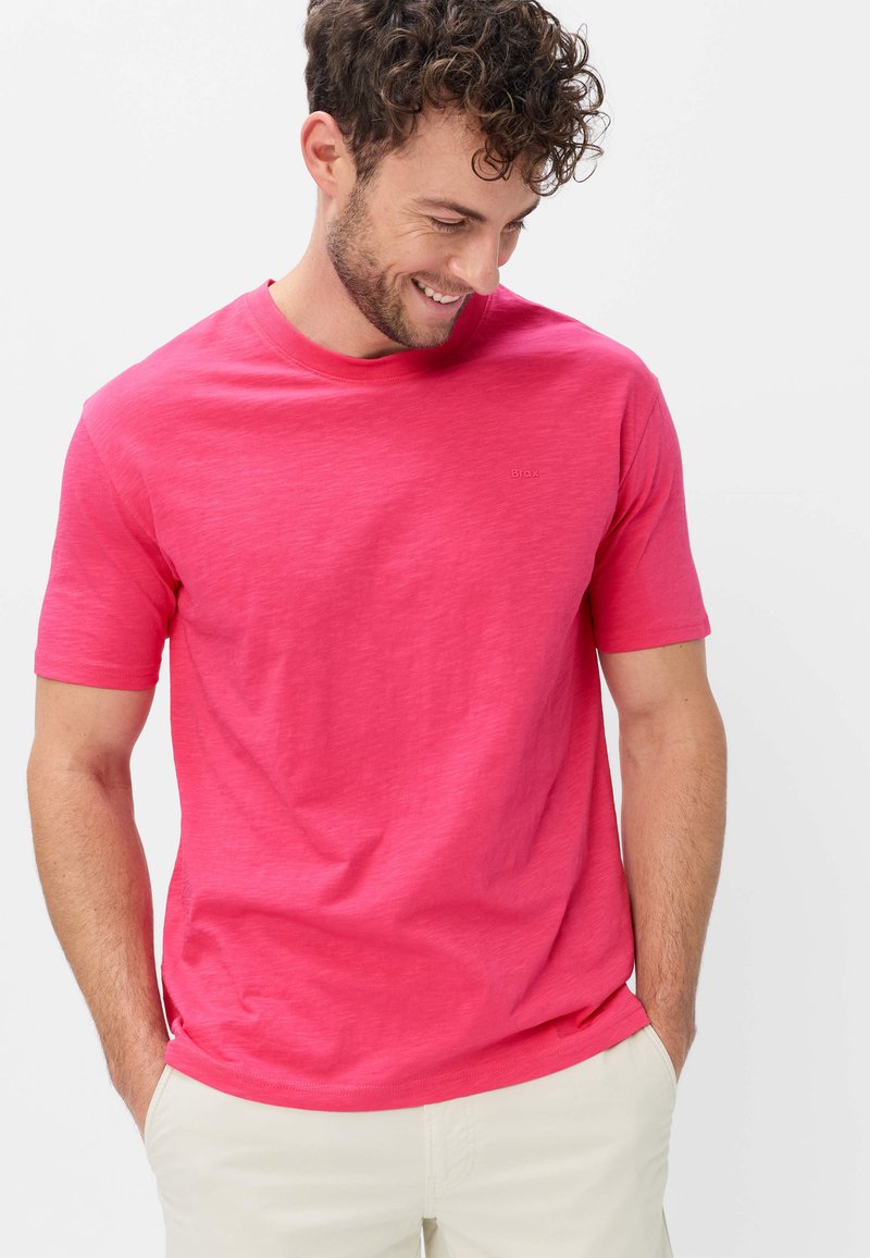 Homme souriant aux cheveux bouclés portant un t-shirt rose vif à manches courtes et un pantalon beige clair, les mains dans les poches, regardant vers le bas.