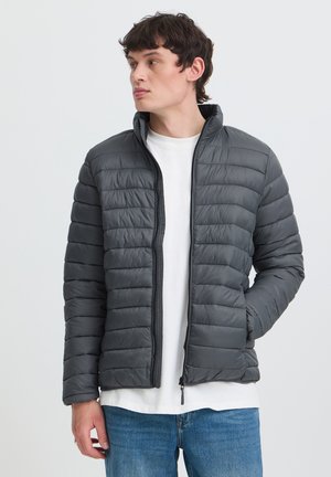 Jeune homme portant une doudoune grise sur un t-shirt blanc et un jean bleu, debout avec une main dans la poche et regardant sur le côté.
