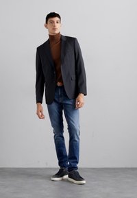 Blazer preto sobre um suéter de gola alta castanho, combinado com jeans azuis e sapatilhas pretas. O conjunto apresenta um corte moderno e um design minimalista.