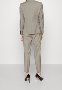 Kvinna i en skräddarsydd beige kostym med korta byxor och svarta lackläder högklackade skor, sedd bakifrån.