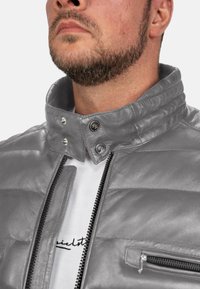 Grijze leren jas met gequiltte textuur, hoge kraag en drukknoopsluitingen. Voorzien van een ritssluiting en een zijzak met een rits.