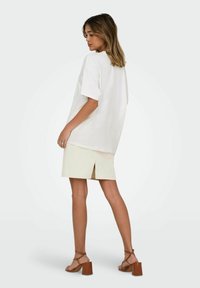 Per didelė balta T-shirt, smėlio spalvos sijonas su skilimu ir rudi aukštakulniai sandalai. Lygus audinys, laisvas kirpimas ir paprastas dizainas.
