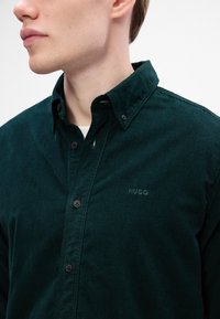 Tume roheline kootud särk nööbiga kraega, mustade nööpide ja rinna peal väikese broderitud "HUGO" logoga.