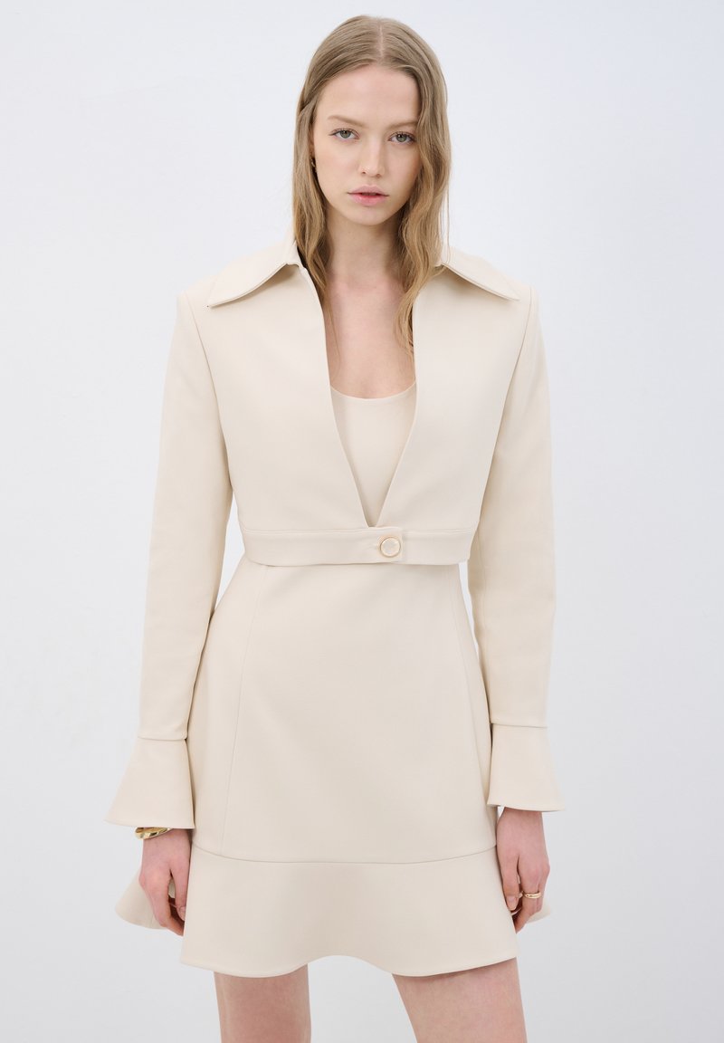 Robe beige en tissu lisse avec un large col, des manches longues et une taille cintrée. Présente un accent de bouton et un ourlet évasé.