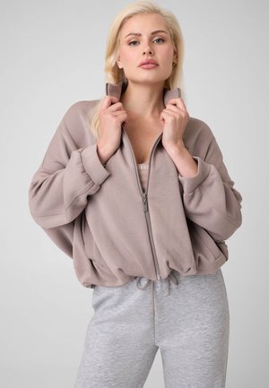 Blonde Frau trägt eine taupefarbene Jacke mit Reißverschluss und hellgraue Jogginghose, hält den Jackenkragen mit beiden Händen vor einem schlichten Hintergrund.