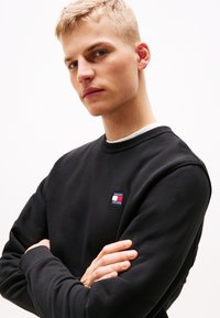 Black sweatshirt with ribbed cuffs and neckline. Features a small logo patch in red, white, and blue on the left chest. Smooth texture.