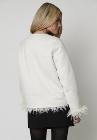Biała kurtka uszyta z gładkiego materiału, z teksturowanymi frędzlami na mankietach, o luźnym kroju i minimalistycznym designie.