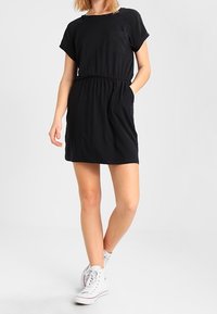Czarna sukienka o krótkim rękawie z okrągłym dekoltem, elastyczną talią i przednią kieszenią; połączona z białymi trampkami na wysokiej podeszwie.