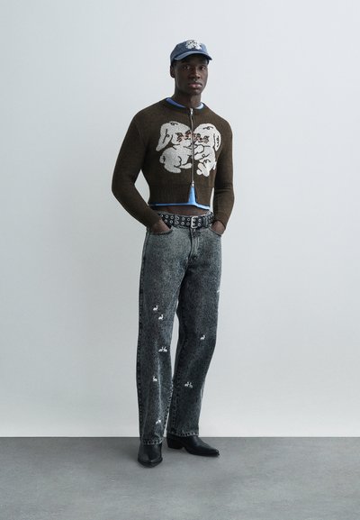 Ung man som bär en brun kort zip-tröja med vitt djurmotiv, syra-tvättade jeans med små vita lappar, svarta stövlar och blå keps.