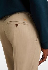 Pantaloni beige con texture liscia, dotati di una tasca posteriore con bottone e un logo di cane ricamato sopra la cucitura della tasca.