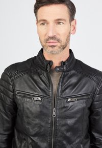 Schwarze Lederjacke mit hohem Kragen, Reißverschluss vorne und zwei Brusttaschen. Verfügt über eine gesteppten Details an den Schultern. Glatte Textur.