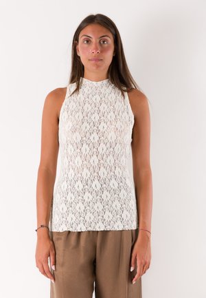 Donna con lunghi capelli castani che indossa un top bianco in pizzo senza maniche e pantaloni marroni larghi, in piedi davanti a uno sfondo chiaro e uniforme.