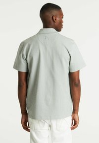 Chemise à manches courtes dans un motif rayé vert clair et blanc. Présente un col décontracté et un logo discret au dos. Texture en mélange de coton.