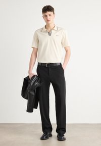 Beige Poloshirt mit gestreiftem Kragen, schwarze Hose und schwarze Lederjacke. Die Schuhe sind schwarz, und ein schwarzer Gürtel vervollständigt das Outfit.