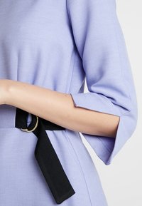 Žena v světle fialových šatech s rukávy do loktů a černým látkovým páskem se zlatou kulatou sponou.