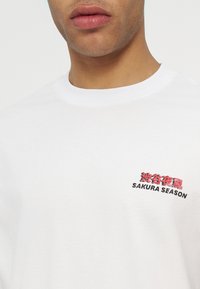 Άνδρας που φοράει λευκό μπλουζάκι με στρογγυλή λαιμόκοψη και κόκκινο και μαύρο κείμενο "SAKURA SEASON" με ιαπωνικούς χαρακτήρες στο αριστερό στήθος.