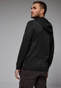 Svart långärmad hoodie med huva och ribbade muddar, gjord av mjukt material. Den har en ren design och subtil brandingdetalj på sidan.