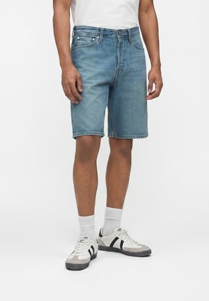 Άνδρας που φοράει μπλε τζιν σορτς μήκους μέχρι τη μέση, λευκές κάλτσες, λευκά και μαύρα αθλητικά παπούτσια και ένα απλό λευκό μπλουζάκι, στέκεται μπροστά σε λευκό φόντο.