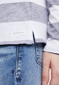 Šedý a bílý pruhovaný tričko s dlouhým rukávem a bočním rozparkem, s malou nášivkou MOPD. V kombinaci s modrými džínami.