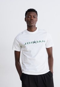 Hvit bomull T-skjorte med "JORDAN" i grønn tekst og en svart Jordan-logo. Korte ermer og rund hals, jevn tekstur.