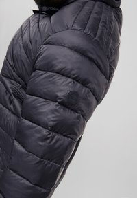 Chaqueta acolchada negra con un diseño acolchado, que presenta un parche con un logo redondo en la manga. El material parece suave y brillante.