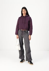 Felpa viola tagliata con cappuccio e cordino, pantaloni grigi a gamba larga e sneakers nere con strisce bianche. Tiene una piccola borsa bianca.