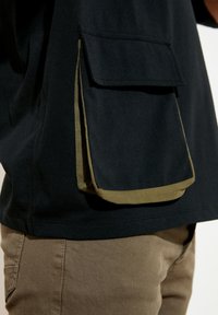 Czarna koszula z dużą kieszenią naszywaną z oliwkowym wykończeniem. Tkanina wydaje się miękka, o gładkiej fakturze i luźnym kroju.