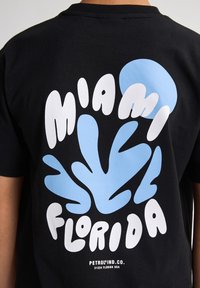 Camiseta de algodón negra con gran texto blanco y azul claro y diseños abstractos que dice "Miami Florida" en la parte posterior.