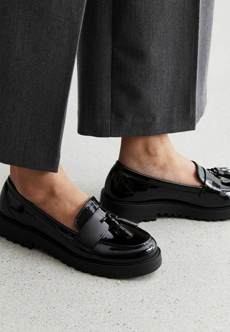 Mocasines de charol negro con una suela gruesa y texturizada, que presentan un detalle decorativo de borlas en la parte superior. Lleva puestos con pantalones grises.