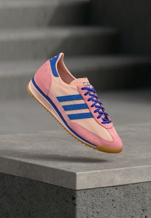 Różowo-beżowy sneaker z niebieskimi paskami i sznurowadłami unoszący się nad betonową krawędzią na rozmytym ciemnym tle.