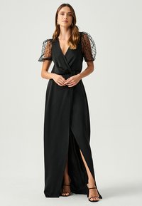 Black wrap dress with polka dot sheer puff sleeves, a deep V-neck, and a side slit. Smooth fabric and cinched waist detail.