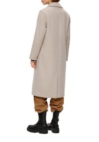Langer beiger Mantel mit einem Reverskragen, aus texturiertem Stoff. Getragen über braunen Hosen und schwarzen kniehohen Stiefeln, mit einer geschlossenen Rückansicht.
