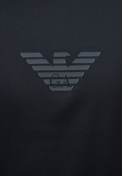 Στυλιζαρισμένο λογότυπο αετού με οριζόντιες ρίγες και τα γράμματα «GA» στο κέντρο, πάνω σε σκουρόχρωμο υφασμάτινο φόντο.