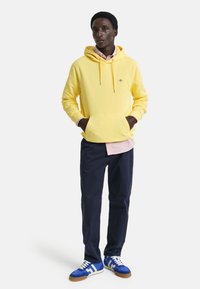 Geel hooded sweatshirt met een voorzak, gecombineerd met een licht gestreept shirt en marineblauwe broek. Afgewerkt met blauwe sneakers met witte accenten.
