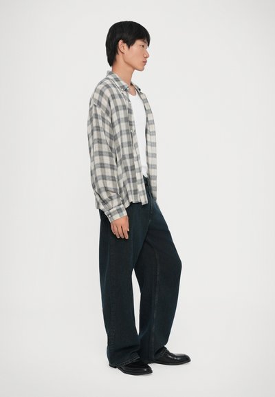 Νέος άνθρωπος με γκρι καρό πουκάμισο πάνω από λευκό μπλουζάκι, φαρδιά σκούρα τζιν και μαύρα παπούτσια, σε προφίλ σε λευκό φόντο.