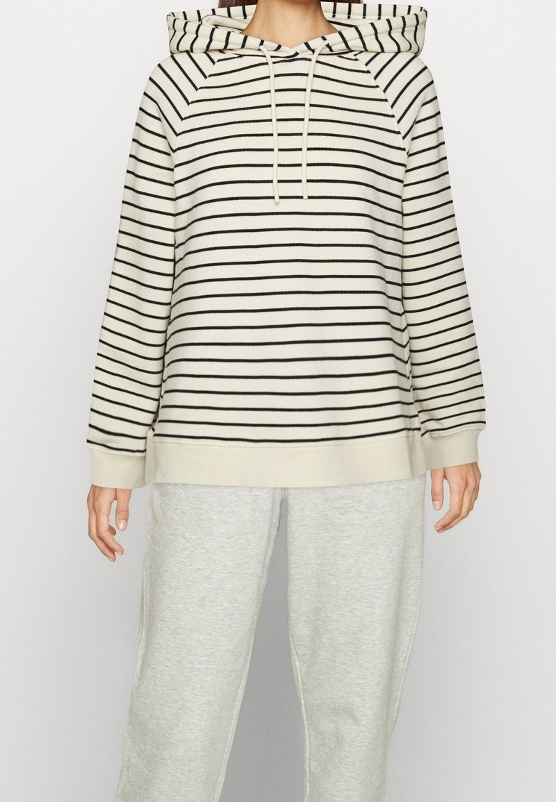 Felpa color crema con strisce nere, dotata di cappuccio con cordino, maniche raglan e polsini a costine, abbinata a pantaloni della tuta grigi chiari.