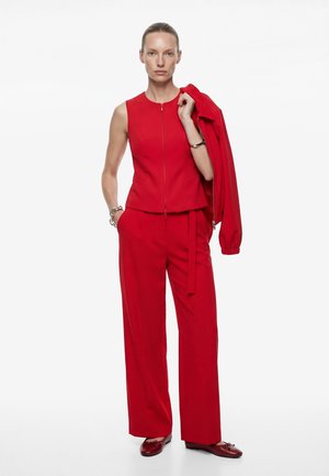 Frau trägt ein ärmelloses rotes Oberteil mit Reißverschluss und weit geschnittene Hose, hält eine passende rote Jacke über der Schulter und steht vor einem weißen Hintergrund.