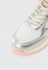 Lightweight running shoe featuring a silver metallic upper, mesh panels, cream laces, and a peach rubber sole. Smooth texture and layered design.