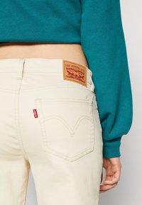 Jeans beige con un acabado texturizado y costuras visibles. Presenta un parche de cuero marrón con logotipo en relieve y una etiqueta roja en el bolsillo trasero.