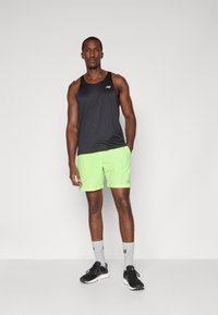 Athletischer Mann in schwarzem ärmellosem Shirt, leuchtend grünen Shorts, grauen Socken und schwarzen Laufschuhen, der vor einem schlichten Hintergrund steht.