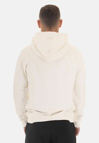 Sudadera con capucha de color crema y mangas largas, con un diseño simple y una textura suave, mostrada desde atrás sobre un fondo liso.