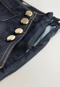 Denim bleu foncé avec boutons dorés et une fermeture éclair partiellement ouverte montrant le tissu en dessous, cousu avec du fil jaune.