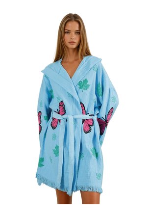 Junge Frau, die einen hellblauen Bademantel mit pinken Schmetterlings- und grünen Blattmustern trägt, steht vor einem weißen Hintergrund.