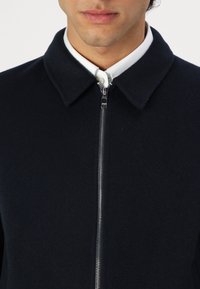 Marineblaue Wollmischungjacke mit spitzem Kragen und frontalem Reißverschluss. Sie verfügt über eine strukturierte Oberfläche und ist über einem weißen Hemd mit Kragen getragen.