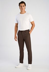 Biała koszulka z krótkim rękawem w połączeniu z ciemnobrązowymi, zwężanymi spodniami. Spodnie mają ściągacz w talii i kieszenie, noszone z białymi trampkami.